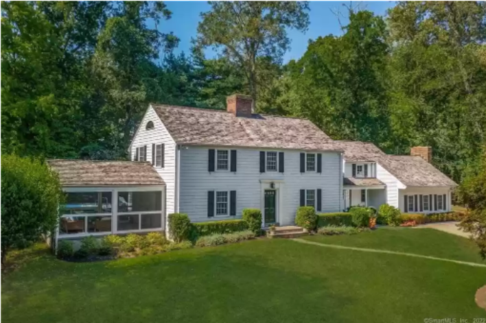 Facade and gray shingle roof of the white Colonial house at 95 Calvary Road, Westport. With foundation plantings, screened porch and lawn, in front of trees and a bright blue sky. 